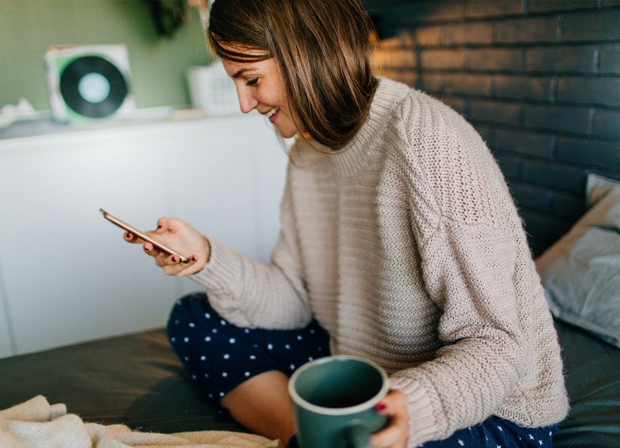 En kvinna sitter på sin säng och bläddrar igenom sin smartphone med en kopp kaffe i andra handen. 