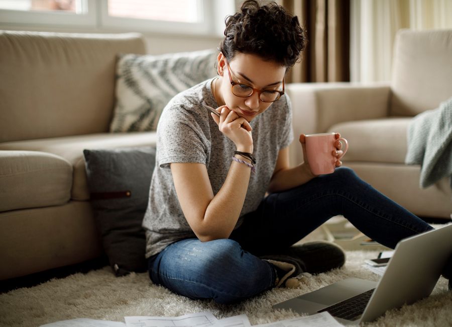 En kvinna sitter på golvet framför sin bärbara dator och håller en penna i ena handen och en kaffekopp i den andra.