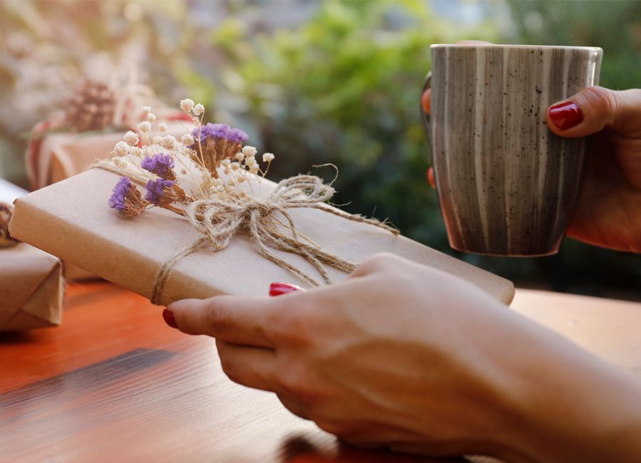 En hand håller en bok som är inslagen i brunt presentpapper och omlindat med ett snöre med små lila och vita blommor fästa vid det. 