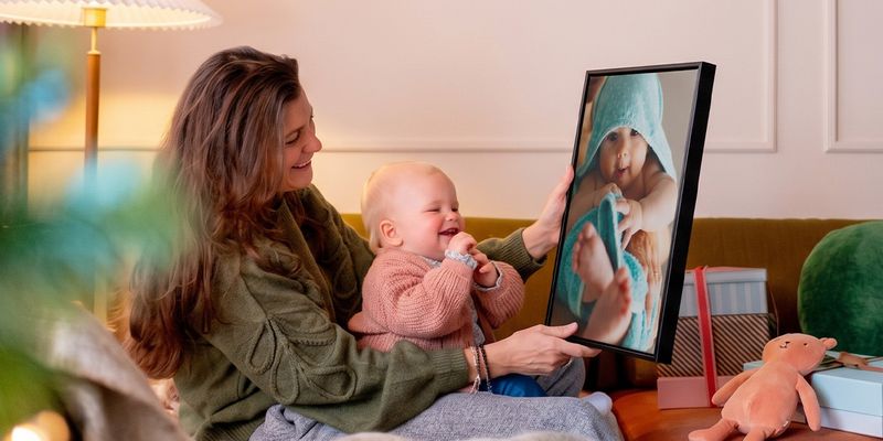 Kvinna med en bebis i knät i ett vardagsrum som håller upp personlig väggkonst med bebisen på, medan bebisen tittar på den och fnittrar, som illustrerar önskefotos guide till fotopresenter för bebisens första jul.