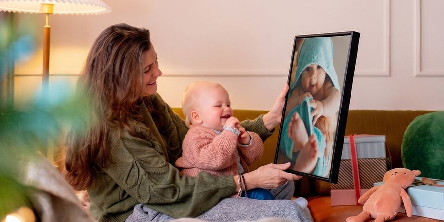 Kvinna med en bebis i knät i ett vardagsrum som håller upp personlig väggkonst med bebisen på, medan bebisen tittar på den och fnittrar, som illustrerar önskefotos guide till fotopresenter för bebisens första jul.