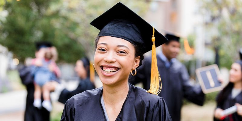 En leende kvinnlig student utomhus i examensklädsel med kåpa och mössa, som inspiration till önskefoto examenspresenter och personliga examensgåvor.