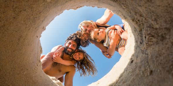 En familj på fyra på stranden – mamma, pappa, dotter och en liten son – tittar ner i en grop som grävts i sanden, och bilden fångar ett lekfullt sommarögonblick som inspirerar till strandbilder och strandfotografering.