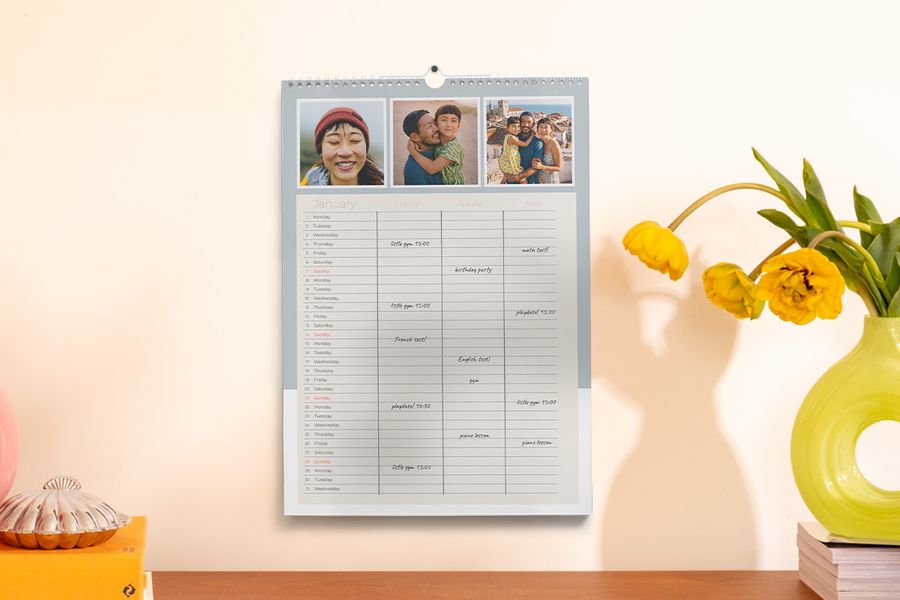 En familjeplanerare hänger på väggen bredvid en vas och några böcker, som illustrerar önskefotos tips om att hålla sig organiserad under skolåret med en personlig planerare.