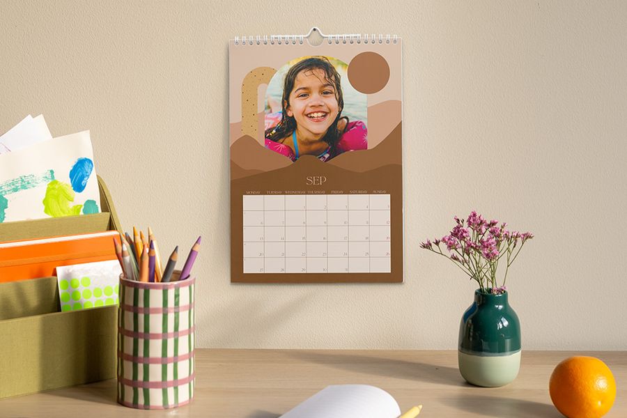 En personlig kalender som visar september och en bild av en ung flicka hänger över ett skrivbord med en pennburk, en vas med blommor och en förvaringslåda med pyssel- och skolmaterial, som illustrerar önskefotos tips om att skapa en fokuserad arbetsplats för skolåret.