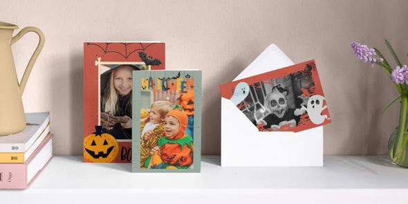 Tre personliga Halloweenkort står sida vid sida på en spiselhylla, med en liten flicka utklädd till häxa, en liten pojke utklädd till pumpa och en liten flicka utklädd till spöke omgiven av spökillustrationer, vilket introducerar önskefotos guide till att skapa grymma Halloween-inbjudningar.
