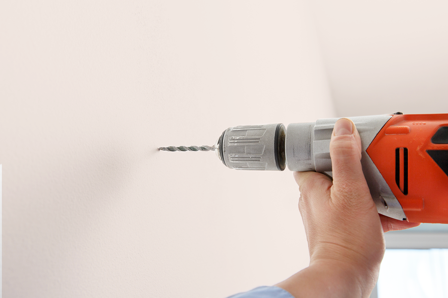 Någon borrar i väggen, vilket illustrerar önskefotos steg-för-steg-tips om att installera hängräls med de medföljande skruvarna och väggpluggen för ett ramupphängningskit.