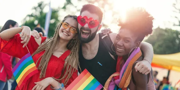 Tre vänner ler på ett Pride-evenemang, håller regnbågsflaggor och bär solglasögon, och firar glädjen och gemenskapen i LGBTQI+ Pride.