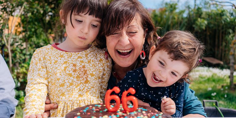 En kvinna ler bredvid sin födelsedagstårta med ljus som visar '66', flankerad av en liten flicka och en liten pojke, vilket introducerar önskefotos ultimata guide till födelsedagspresenter.