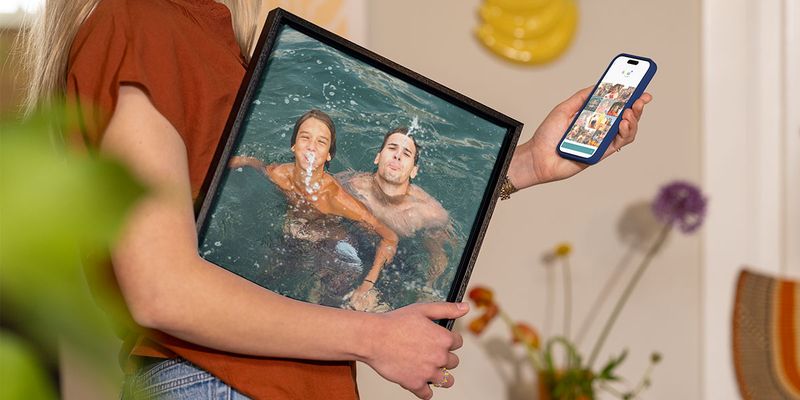En kvinna håller en inramad semesterbild av en man och en liten pojke som leker i vattnet under armen, medan hon väljer ut fler bilder i telefonen, vilket illustrerar önskefotos guide till att göra semesterbilder till väggkonst.