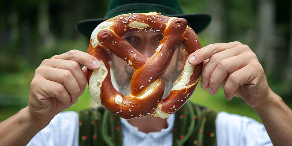 En man i traditionell bayersk klädsel håller upp en stor pretzel framför ansiktet, vilket introducerar önskefotos guide till de bästa Oktoberfest-städerna i Europa.