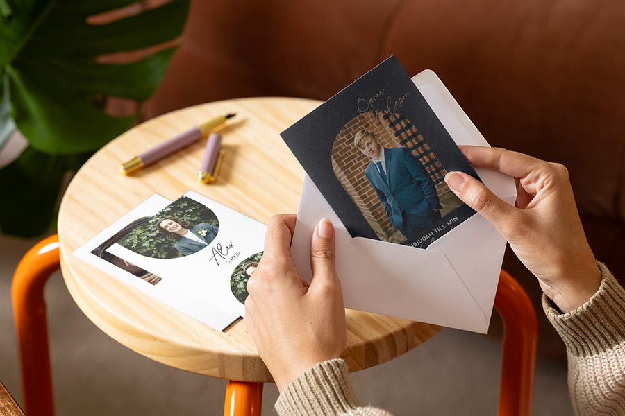 En person som tittar på inbjudningskort till första heliga kommunionen för pojkar från önskefoto, med fler inbjudningar på ett bord i bakgrunden.