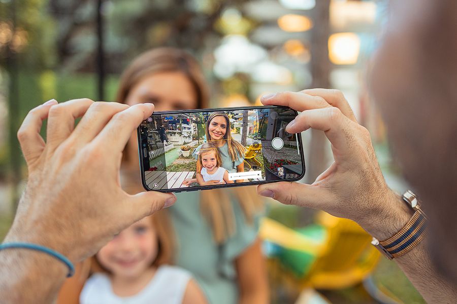 En hand håller en mobiltelefon som fångar en leende mamma och hennes dotter i starkt solljus, och visar hur man tar bilder i starkt solljus med mobilen.