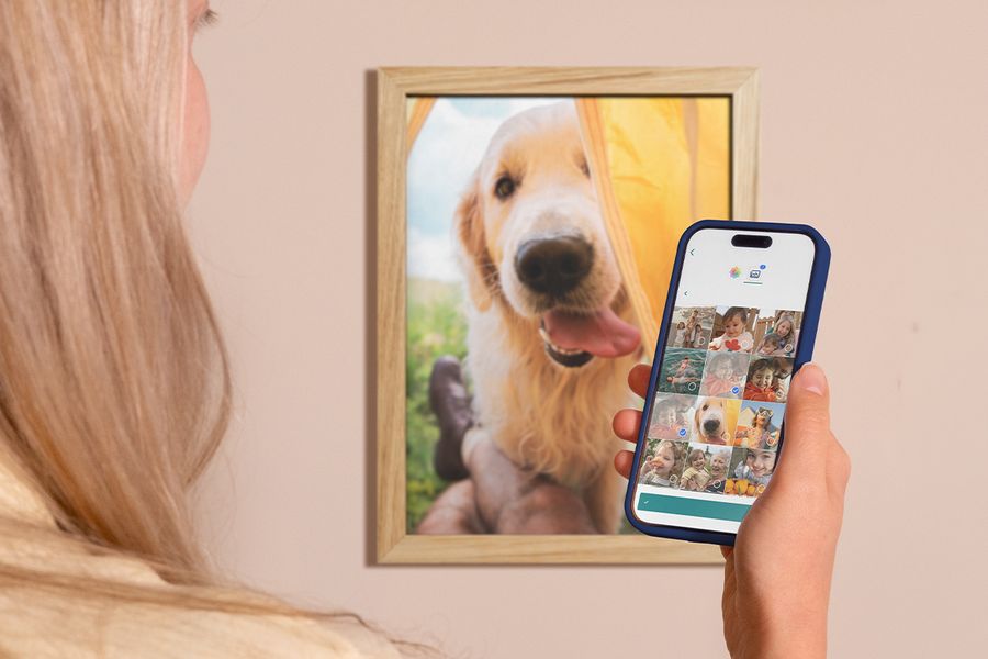 En kvinna tittar igenom kamerarullen på sin mobil, med en inramad bild av en labrador på väggen bakom henne, som inspiration för att organisera digitala foton och välja ut minnen att framkalla.