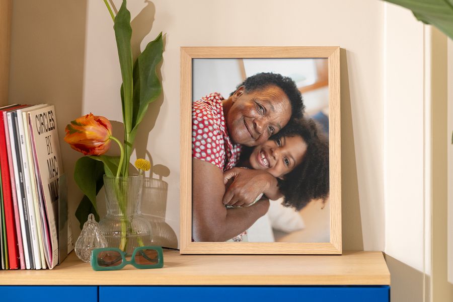 Ett inramat foto av en mormor eller farmor och hennes barnbarn som kramas står på ett bord bredvid böcker, ett magasin och en dekorativ vas, som inspiration till ramtrender och prisvärda sätt att lyfta din väggdekor.