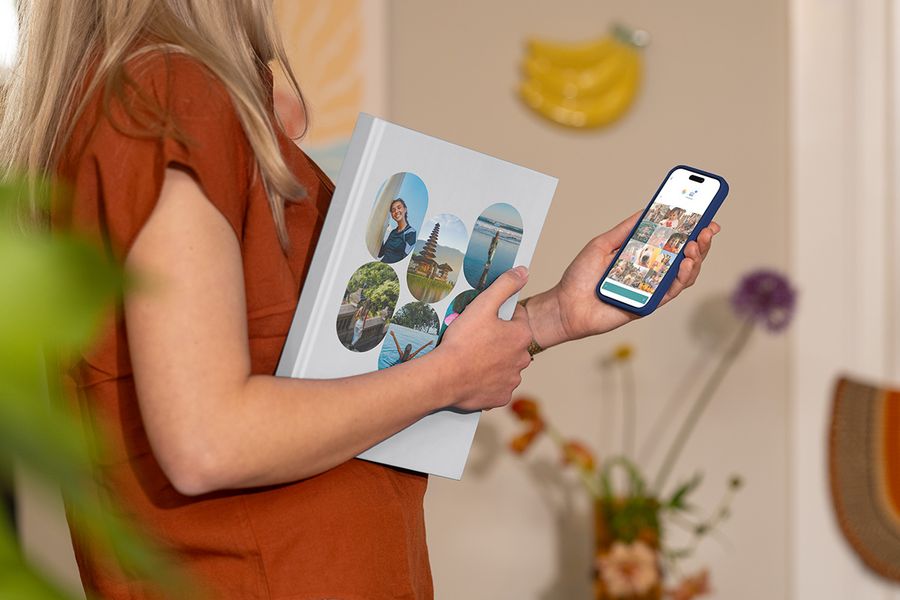 En kvinna håller ett stängt önskefoto semesteralbum med familjens resebilder på omslaget samtidigt som hon tittar på resebilder i mobilen, vilket illustrerar hur man väljer rätt bilder till en personlig fotobok resa.