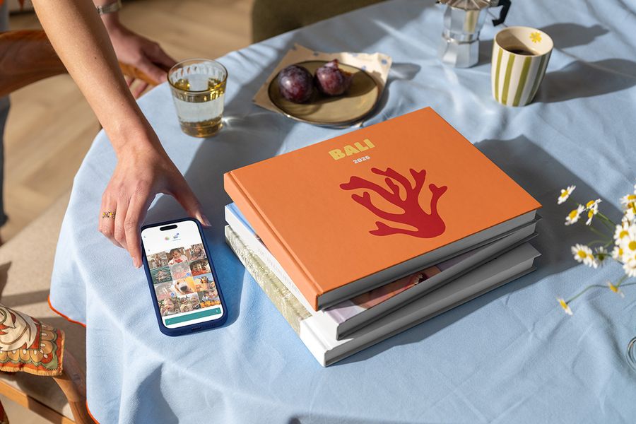 En stapel med olika önskefoto resefotoböcker står bredvid en hand som håller en mobil med en semesterbild redo att använda, med en Bali 2026-bok med korall på omslaget högst upp, vilket visar hur man skapar en personlig fotobok resa.