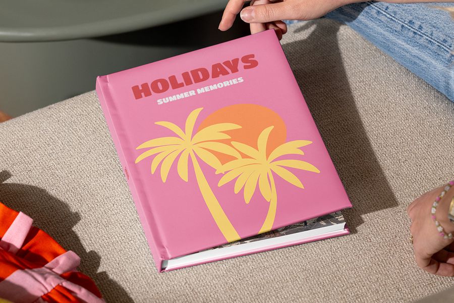 En stängd önskefoto fotobok resa med titeln ”Holidays, summer memories” och illustrerade palmer på ett rosa omslag hålls av händer som är redo att öppna den, vilket inspirerar till hur man strukturerar ett semesteralbum och berättar sin resehistoria.