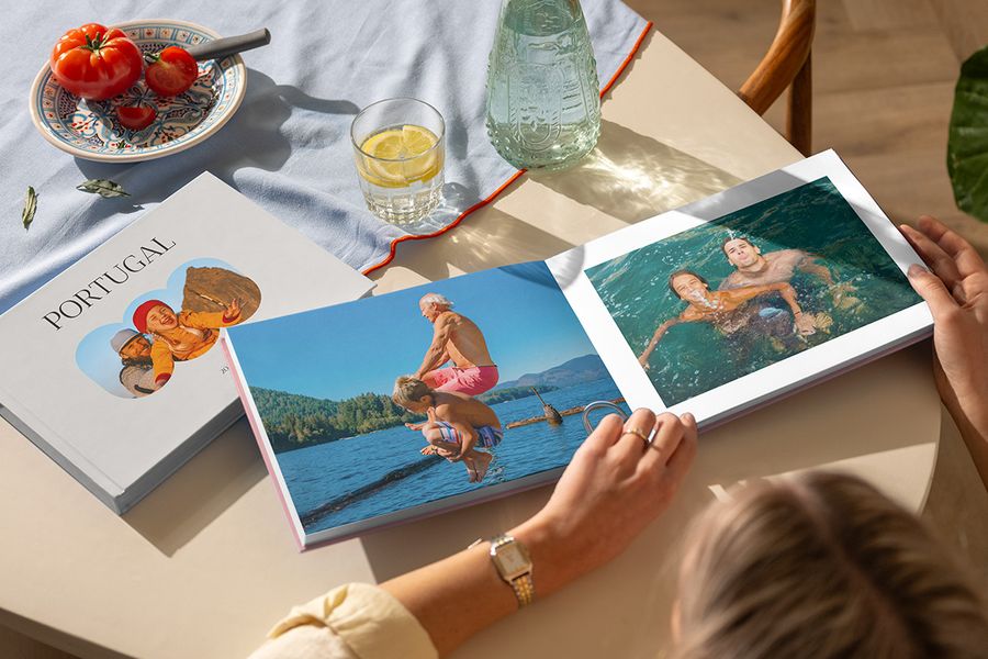 Två olika önskefoto resefotoböcker visas, där en kvinna bläddrar i en öppen fotobok resa med familjebilder från havet, bredvid en stängd Portugal-bok, som illustrerar ett personligt semesteralbum fullt av reseminnen.