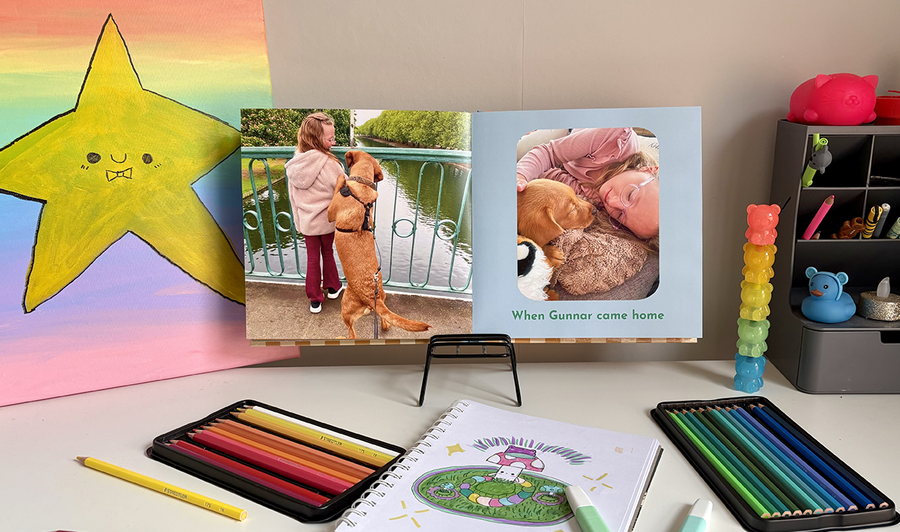 En öppen barnfotobok ligger på ett barns skrivbord omgiven av färgpennor och konstverk, med en bild av en flicka och hennes hund på vänstersidan och en annan bild med bildtext på högersidan, som illustrerar önskefotos tips för att blanda foton och konstverk i en familjefotobok.