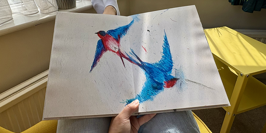 En öppen barnfotobok med ett helsidesuppslag av en fågelteckning ligger i knät på en kvinna som sitter i en fåtölj, som illustrerar önskefotos steg för att skapa en familjefotobok av barnets teckningar.