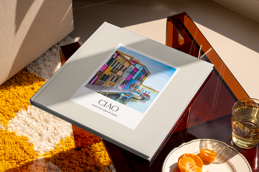 En fotobok med en kanal och färgglada hus i Italien på omslaget heter 'Ciao from the Land of Pasta', som illustrerar önskefotos fotobok titel idéer för stadsresor.