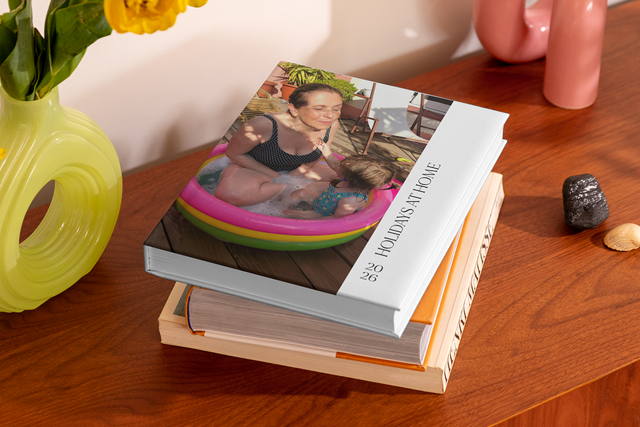 En stapel fotoböcker har högst upp en bok som visar en kvinna och ett litet barn i en pool i trädgården, med titeln 'Holidays at Home', som illustrerar önskefotos fotobok titel idéer för staycation.