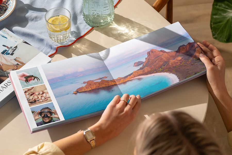 En öppen resefotobok på ett bord visar ett dubbelsidigt uppslag med ett vackert strandlandskap tillsammans med tre sommarbilder, som illustrerar önskefotos Travel Chapter-design från den nya Summer Travel Series.