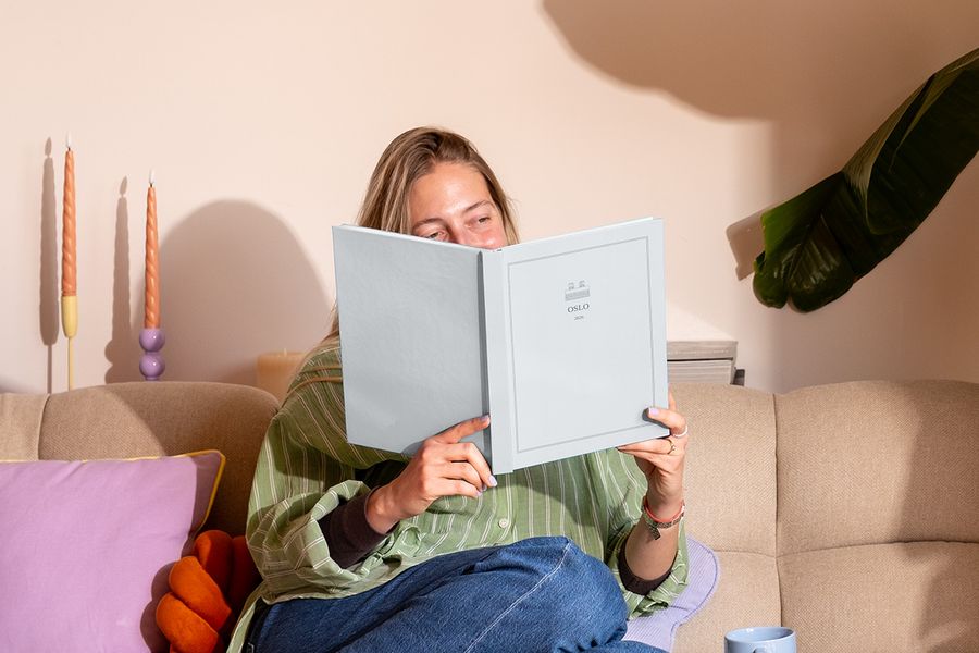 En kvinna sitter i soffan bredvid en färgglad kudde och tittar på en personlig resefotobok med titeln 'Oslo', som visar önskefotos nya Summer Travel Series-design.