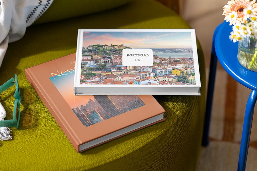 Två personliga fotoböcker ligger staplade på en pall, den översta med titeln 'Portugal' och en bild av Portugal, och under den en bok med titeln 'New York' och en bild av staden, som visar önskefotos personaliserbara Summer Travel Series-design.