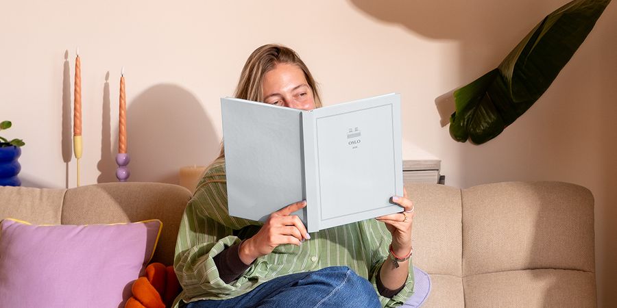 En kvinna sitter i soffan bredvid en färgglad kudde och tittar på en personlig resefotobok med titeln 'Oslo', som visar önskefotos nya Summer Travel Series-design.