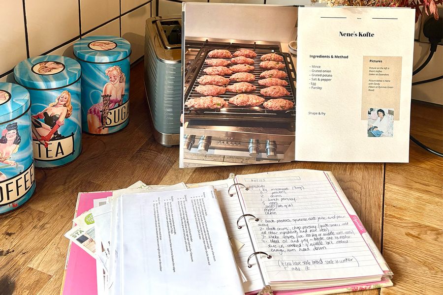 Sarahs öppna personliga kokbok visar ett recept på hennes Nenes kofte, placerad framför en gammal pärm öppnad för att visa handskrivna familjerecept, båda liggande på en köksbänk, vilket illustrerar hur hon samlade generationers recept i en bok.