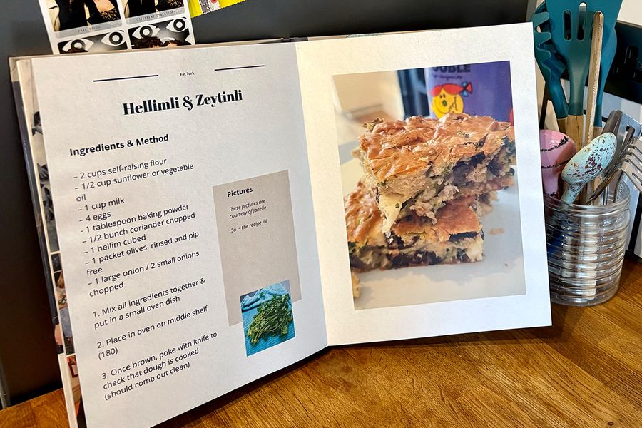 En öppen personlig kokbok ligger i ett kök, med en burk med skedar och köksredskap i bakgrunden, vilket illustrerar önskefotos tips för att välja vilka recept som ska inkluderas innan du skapar din egen kokbok.