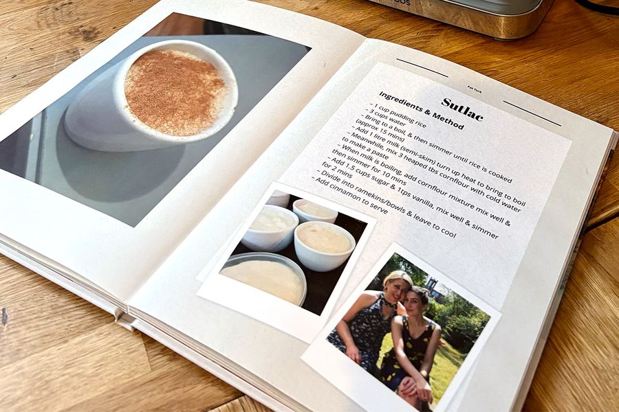 Sarahs öppna receptbok ligger på köksbänken och visar sitt layflat-fotoboksformat, med bilder av turkisk risgrynsgröt på vänstersidan och familjens motsvarande recept på högersidan, vilket illustrerar önskefotos tips om att välja en layflat-design som är lätt att laga mat från.