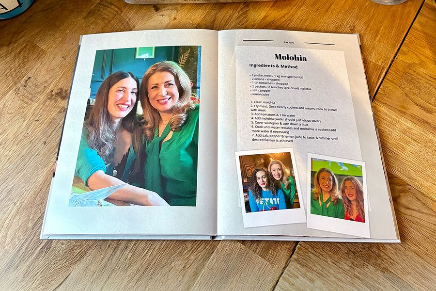 Ett uppslag från Sarahs personliga receptbok visar ett recept på molohia, tillsammans med familjebilder istället för en bild av rätten, vilket illustrerar önskefotos tips om att inkludera personliga bilder som gör en receptbok verkligt meningsfull.