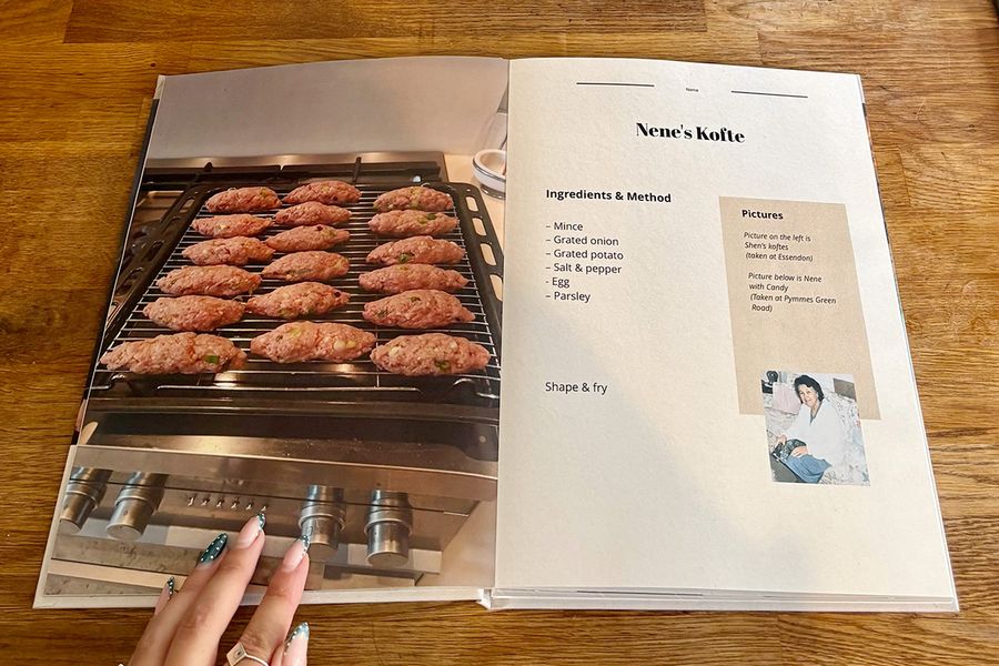 Ett öppet uppslag från Sarahs receptbok visar hennes Nenes kofte-recept, med en manikyrerad kvinnohand vilande på boken på en träbänk, vilket illustrerar önskefotos sista checklista-tips innan man skriver ut en personlig kokbok.