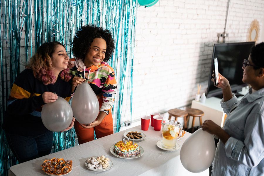 Två flickor poserar framför en glittrig fotobakgrund, håller ballonger bredvid ett snacksbord fullt med festmat och en födelsedagstårta, vilket illustrerar önskefotos tips för att fotografera festdekor och matupplägg.