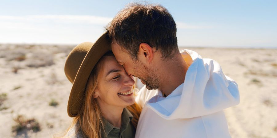 Ett par i trettioårsåldern ler och kramar varandra i en strandaktig, ökenliknande miljö, vilket introducerar önskefotos guide med 60 idéer till meddelanden i ett bröllopsdagskort.