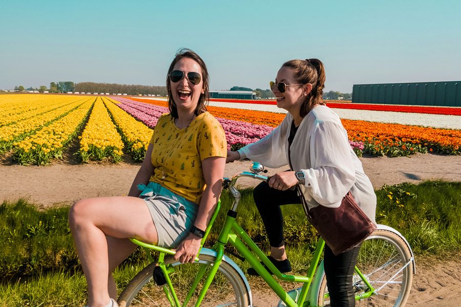 Två kvinnor cyklar tillsammans, ler och skrattar i ett blomsterfält, vilket illustrerar önskefotos idéer för vad man kan skriva i ett bröllopsdagskort till vänner.