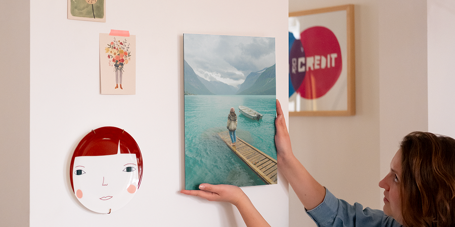 En kvinna sätter upp personlig väggkonst på sin vägg, bredvid en dekorativ tallrik och några fotoutskrifter, vilket introducerar önskefotos steg-för-steg-guide för att hänga upp väggkonst med upphängningskit.