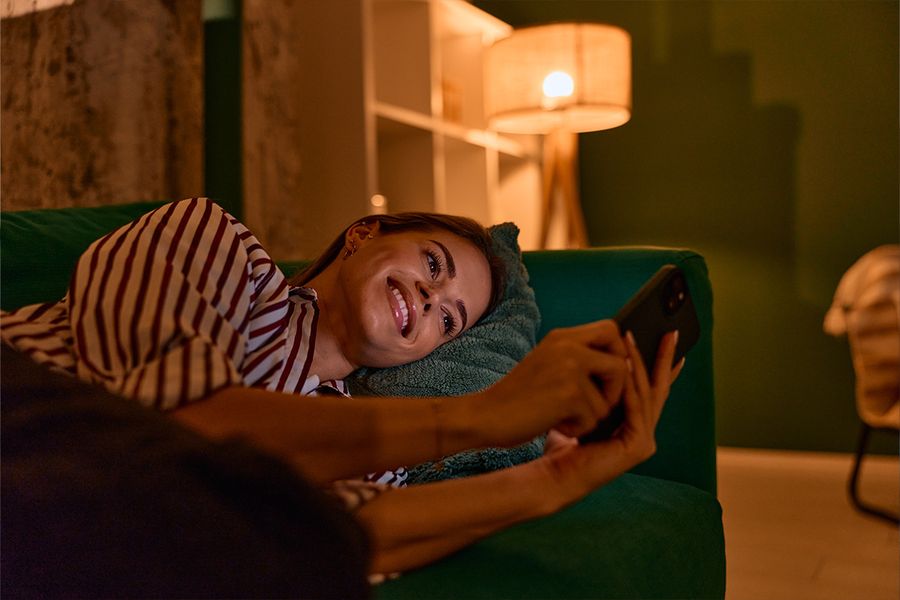 En kvinna ligger på en mörkgrön soffa och tittar på sin telefon i ett vardagsrum, upplyst av en lampa för mysig belysning, vilket illustrerar önskefotos tips för hygge-belysning hemma.