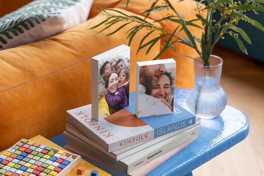 Två personliga fotoblock vilar på ett urval soffbordsböcker på ett blått sidobord bredvid en orange sammetssoffa, vilket illustrerar hur önskefotos fotoblock ger en personlig touch åt ett hygge-hem.