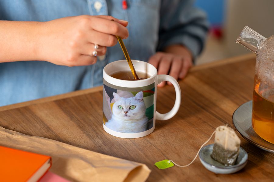 Någon rör om med en sked i en personlig fotomugg med en bild av en vit katt medan de gör en kopp te, vilket illustrerar önskefotos tips om att avsluta en hygge-läshörna med en personlig mugg.