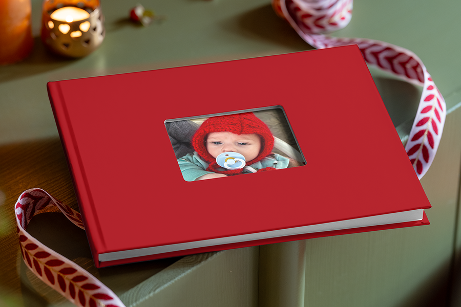 Röd fotobok med ett utskuret fönster på omslaget, där man ser en nyfödd bebis med en röd stickad mössa och en napp, som illustrerar önskefotos tips för att skapa en fotobok med bebisens första minnen.