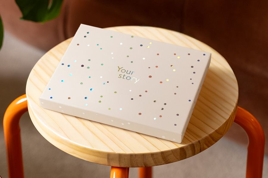 En önskefoto presentationsbox står på en träpall, dekorerad med silverfärgade holografiska cirklar och texten 'Your Story', vilket illustrerar den matta elfenbensfärgade designen med en touch av holografisk lyster.
