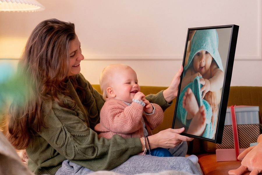 Kvinna med en bebis i knät i ett vardagsrum som håller upp personlig väggkonst med bebisen på, medan bebisen tittar på den och fnittrar, som illustrerar önskefotos idéer för att göra unik väggkonst av bebisbilder till bebisens första jul.
