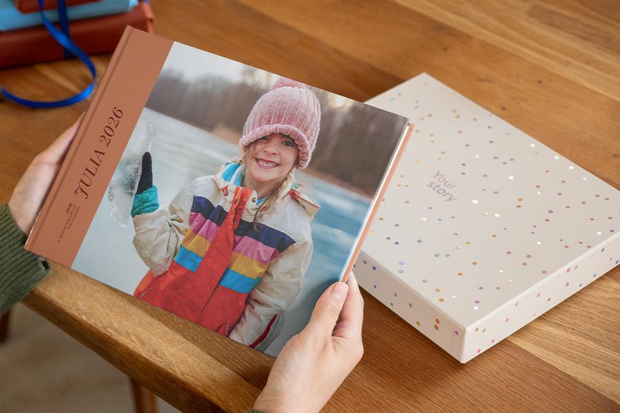 Någon håller i en fotobok med titeln 'Julia', med en leende flicka på omslaget, med en presentationsgåvobox liggande precis bakom på bordet, vilket illustrerar varför en fotoboksbox gör en skapelse extra speciell.