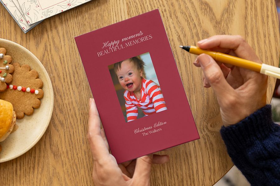 Händer redo att signera ett personligt julkort med en leende bebis, en julhälsning och ett familjenamn på framsidan, bredvid en tallrik med julkakor på ett bord, som illustrerar önskefotos idéer för personliga julkort till bebisens första jul.
