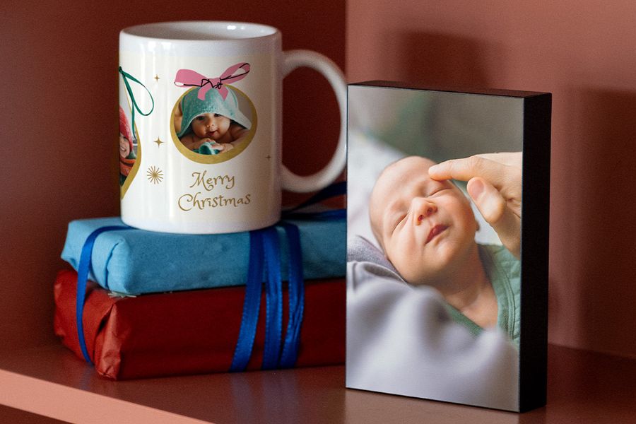 Personlig mugg med en bild på en bebis i en handduk och en god jul-hälsning, placerad ovanpå två inslagna paket bredvid ett fotoblock med en bild på en sovande nyfödd, som illustrerar önskefotos idéer för fotopresenter till nyblivna föräldrars första jul.