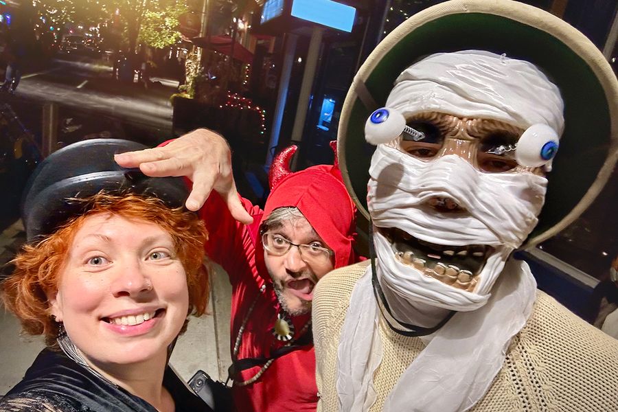 Tre vuxna i Halloween-kostymer på gatan, en med skelettmask med utstickande ögon, en utklädd till djävul och en kvinna med svart hatt, vilket illustrerar önskefotos Halloween-citat för maskeradkläder.